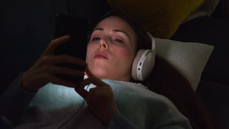 Mujer-Con-Auriculares-Inalámbricos-Tumbada-En-El-Sofá-De-Casa-Por-La-Noche-Transmitiendo-Música-O-Viendo-Una-Película-En-Un-Teléfono-Móvil-4